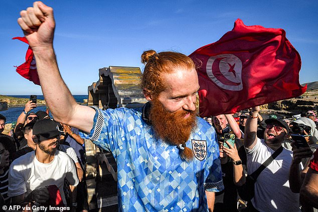 A man holds up a Tunisian national flag behind runner Russ Cook as the latter gestures by the memorial sign marking the northern-most point of Africa upon arrival at Cape Angela