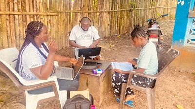 A man,a woman and a yuoung person sit on plastic chairs in front of a small table, in a dry yard. The man and the woman have laptops open.