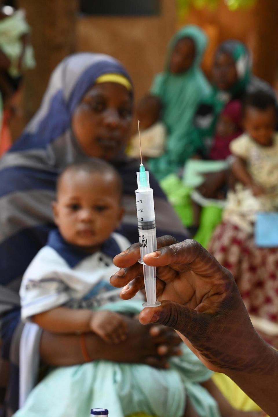 A woman and her baby look at a hand holding a syringe