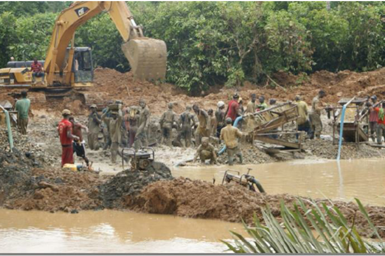 A yellow excavator dwarfing several men perched on higher ground, mechanical scoop poised above mining pit filled with dirty brown water in Ghana. Image by Danyo Gilbert.