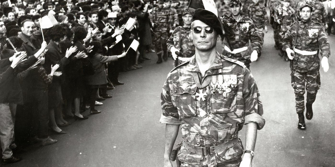 A military officer in sunglasses and a beret leads a group of soldiers behind him through a crowd