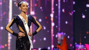 Miss USA contestant with medium-brown skin, wearing a dress with cutouts on the arms, stomach and chest, wearing a USA sash and smiling toward the camera. She stands in front of a purple sparkly backdrop. People in the foreground hold up cameraphones