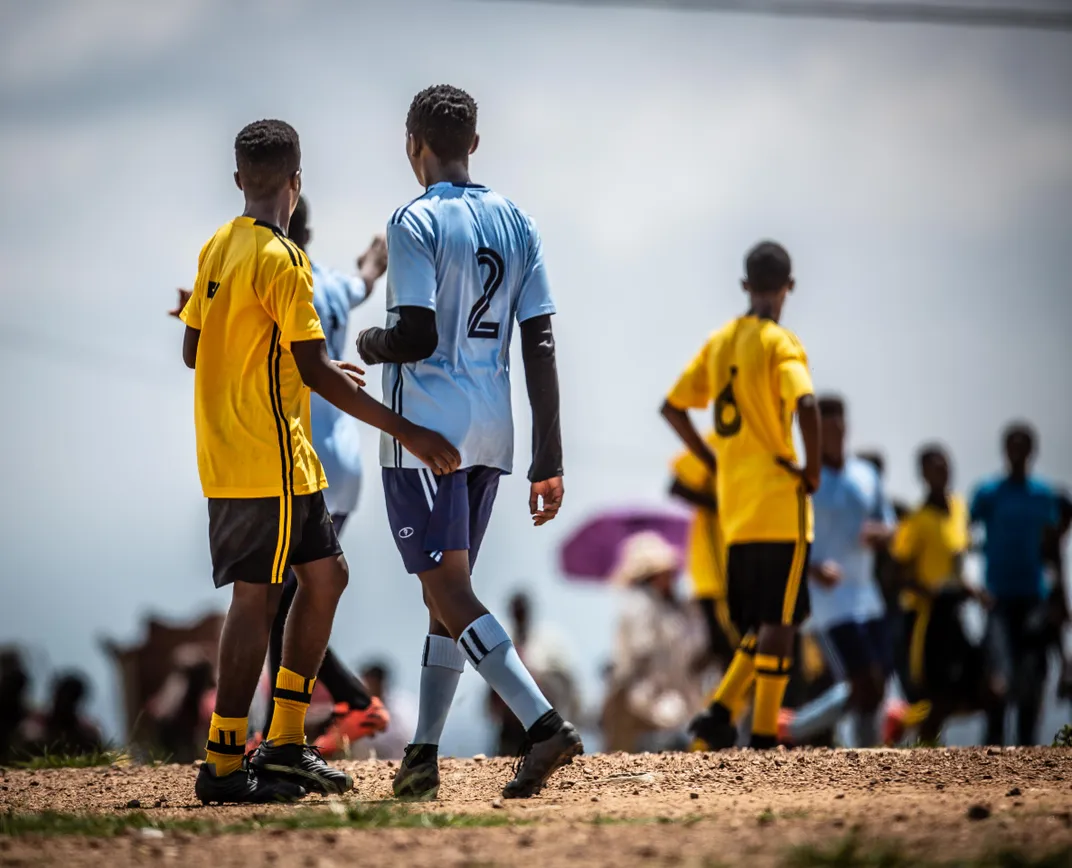 8 - In a rural community, teenagers gather to play soccer among cheering supporters.