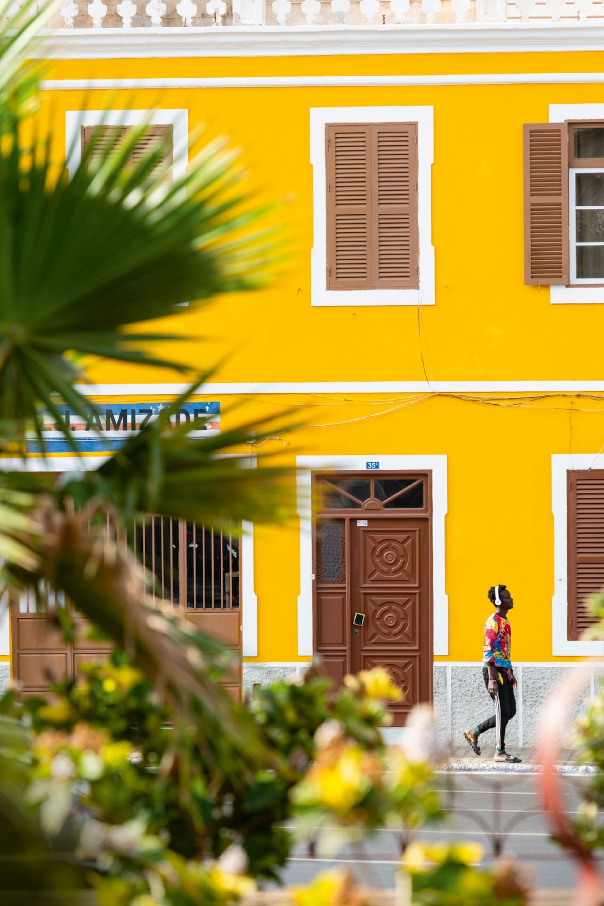 Man wearing headphones walks in front of a brightly painted yellow building with shutters