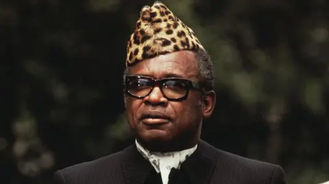 Getty Images Former President of the Democratic Republic of Congo Mobutu Sese Seko wearing a leopard-skin hat, glasses and a dark suit and cravat